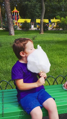 Joyful little boy in casual purple outfit relaxing on bench after active ride. Smart kid enjoying sweet sugar dessert in green park nature
