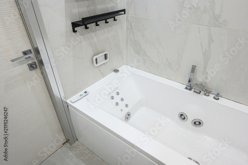 Modern compact bathroom features white jacuzzi tub with jets, black wall shelf, marble tiles, chrome fixtures, handheld showerhead, control panel, minimalist design