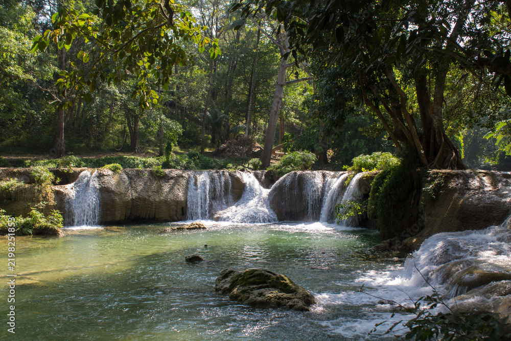Fototapeta premium Tropical jungle waterfall in Thailand surrounded by lush green rainforest, moss-covered rocks, dense vegetation, and flowing water. The natural cascade creates a peaceful and refreshing scene