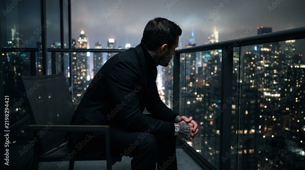 Fototapeta premium Serious businessman sitting alone on a city balcony at night with smartphone