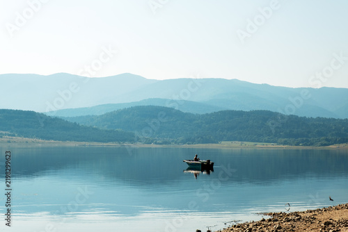Fisherman In Small Boat Fishing Calm Lake Surface Blue Mountain Layers Sky Early Morning Recreation Hobby Freedom Batak Reservoir Bulgaria Nature Scenic Stock Photo