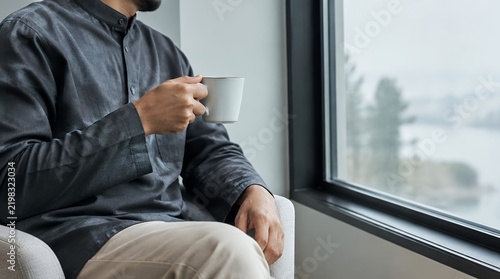 Adult Muslim man in dark attire reading book while gazing out window, realistic commercial photo