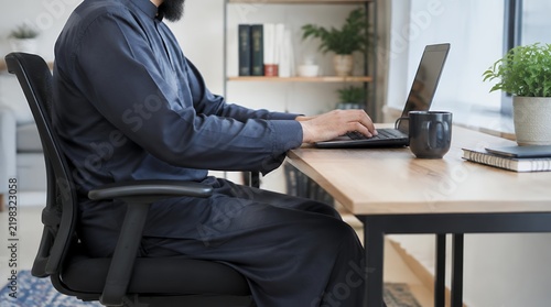 A Muslim man wearing a blue thobe and black face mask works intently on a laptop at his home office desk in 2026