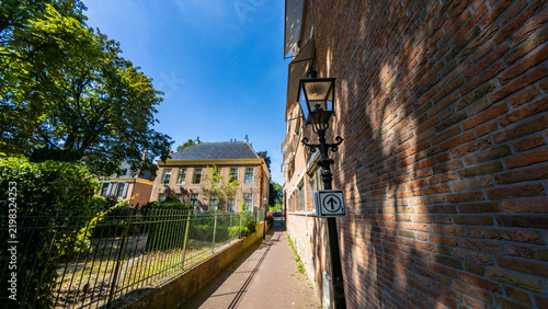 A narrow paved alleyway flanked by a traditional brick building and an old street lamp, with historic buildings and trees in the background in the Netherlands.