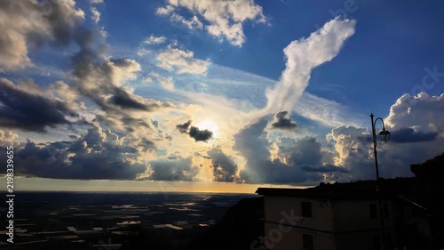 Fast moving clouds reveal radiant sun rays over a vast open plain, framed by a silhouetted house and streetlamp. Dynamic skies inspire a deep sense of natural beauty and atmospheric evolution.