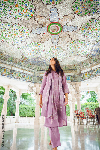 Portrait of a young Indian woman in traditional dress standing at an ancient temple in India, with carved stone pillars softly blurred in the background.