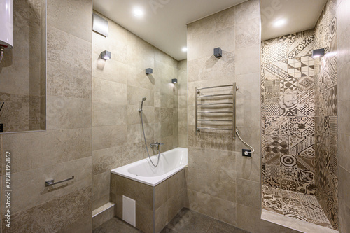 Modern bathroom with beige stone tiles, geometric patterned accent wall, built-in bathtub, showerhead, towel warmer, recessed ceiling lights, wall sconces, niche, grab bar