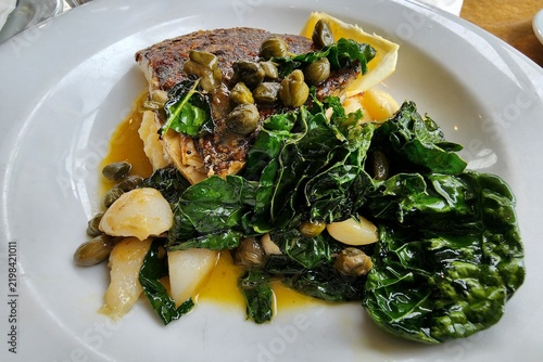 Photography Sydney, Australia - Gourmet pan‑seared fish with crispy skin served on white plate with capers, sautéed greens and tender onions in light sauce under warm restaurant lighting