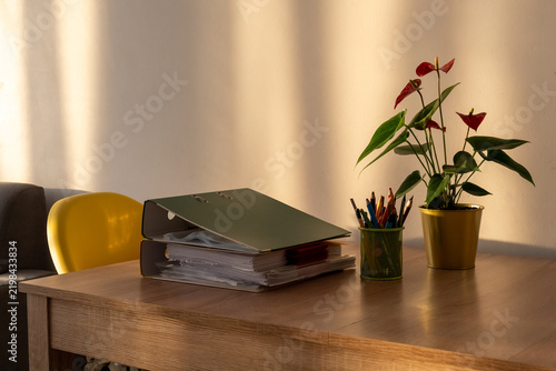 Office desk with stacked binders, colored pencils and red anthurium beside yellow chair. Concept of school administration, educational paperwork, lesson preparation and academic organization.