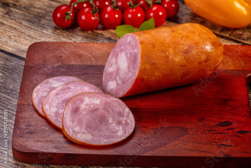 Sliced ham sausage on dark wooden cutting board with cherry tomatoes and yellow pepper in background, rustic food presentation