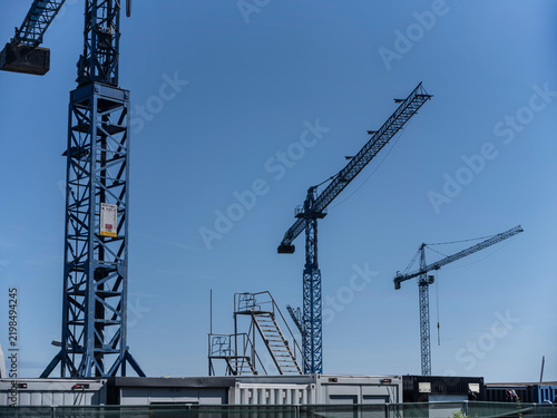 Foto Blue lattice tower cranes rise above site cabins and fencing against a clear blue sky, a bright image of construction and heavy industry