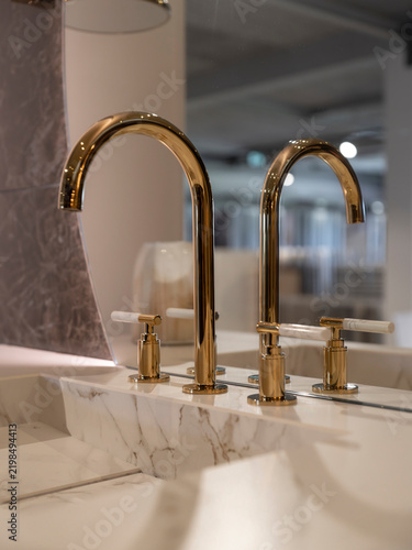 Vertical close-up of a classic gold three-hole faucet on a white marble basin, mirrored at the edge, with dark marble and soft glow behind.