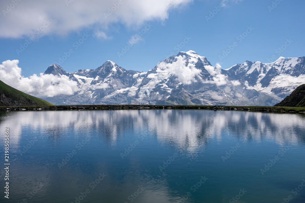 Fototapeta premium Dramatic snow-covered mountain range mirrored in calm blue alpine water beneath a clear summer sky.