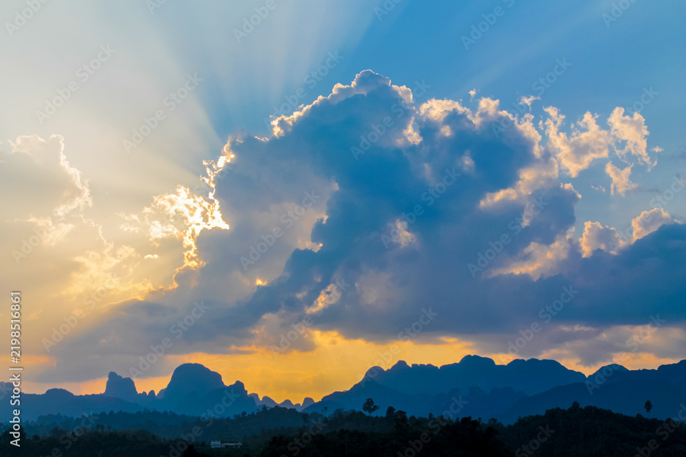 Obraz premium Dramatic tropical sunset over the seaside, with golden sun rays breaking through layers of clouds and illuminating the sky above the calm ocean. The warm evening light creates a striking atmosphere, w