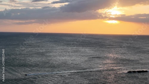 A boat moves through the ocean as the sun sets on the horizon. The sky shows warm colors while waves roll in.