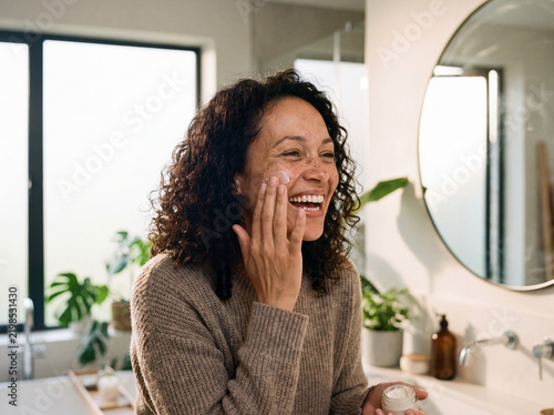 Smiling mature woman applying facial moisturizer cream in a bright modern bathroom, embracing natural skin texture and daily self-care routine