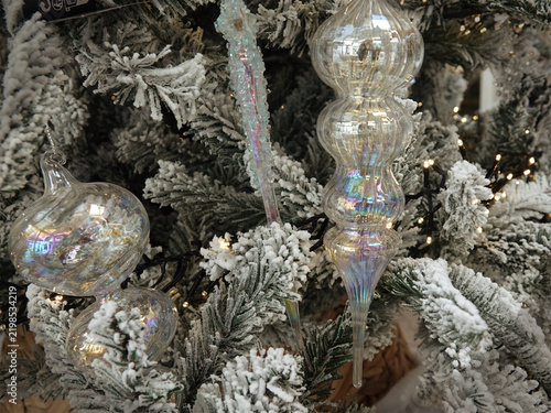 Snow-covered Christmas tree branches adorned with iridescent glass ornaments. The decorations include a round, bulbous one and an elongated, spiral icicle shape. 
