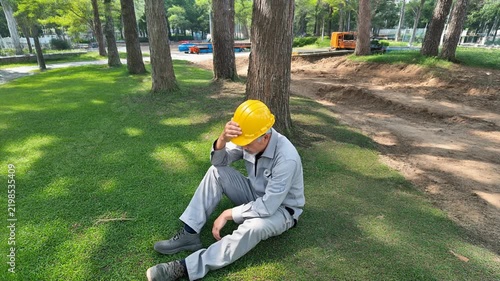 黄色い安全ヘルメットの中年男性作業員が木陰の地面に座り額を覆って疲労を休める