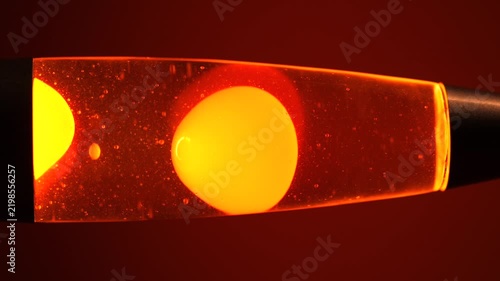 Close Up of Retro Lava Lamp with Rising and Falling Yellow Wax Blobs, Warm Orange Ambient Light and Red Background