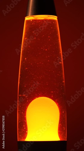 Close Up of Retro Lava Lamp with Rising and Falling Yellow Wax Blobs, Warm Orange Ambient Light and Red Background