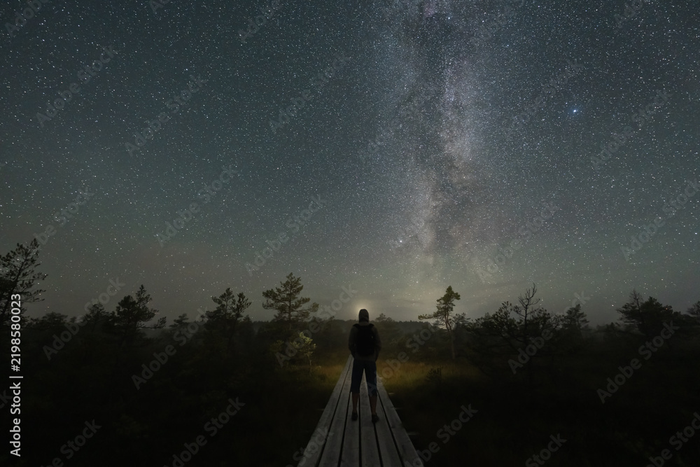 Fototapeta premium Man standing on wooden boardwalk in dark bog facing starry sky with Milky Way galaxy in Estonia nature reserve during clear night.