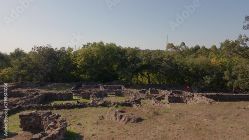 An elevated aerial perspective showcases the stone foundations of an ancient Iron Age settlement surrounded by lush green trees under a clear sky.