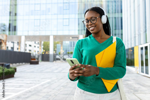 Smiling black student listening music using smartphone and headphones outdoors