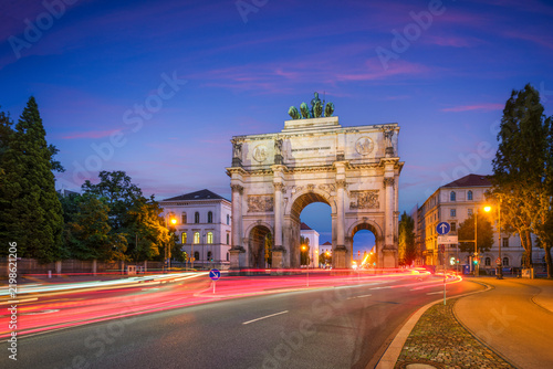 Dramatic twilight view of the Siegestor monument in Munich, Bavaria, Germany, featuring vibrant red and white car light trails on the road.