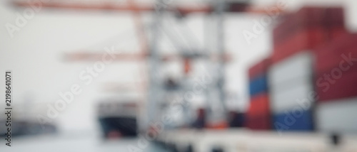 A blurry image showcasing a busy shipping port with cranes and container ships. The colors of the containers range from red to blue, emphasizing maritime activity.