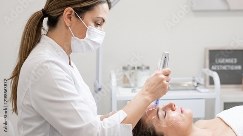 Female cosmetologist in face mask, with silver microneedling pen over forehead gel, performing skin rejuvenation treatment, modern aesthetic clinic room, cosmetology photography