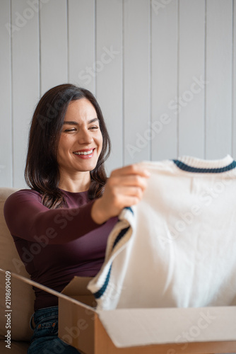 Smiling woman holding up a knitted sweater from an open delivery box, sitting on a sofa. Female customer checking an online shopping purchase