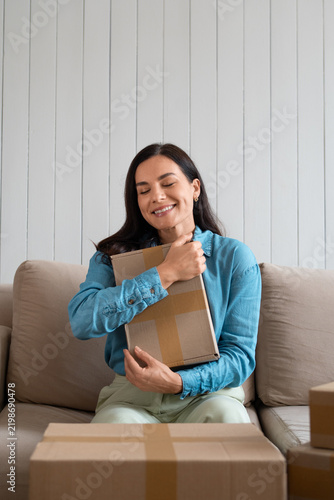 Happy woman hugging a cardboard parcel, eyes closed, surrounded by delivery boxes on the couch. Female customer celebrating a purchase