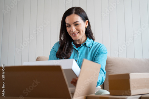 Smiling woman lifting a white box from an open parcel, more boxes nearby on the couch. Female customer unboxing deliveries