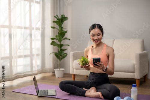 Asian woman eating salad, using phone on yoga mat
