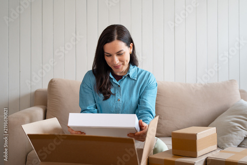 Smiling woman holding a white product box beside stacked parcels on the couch. Female customer checking an online purchase
