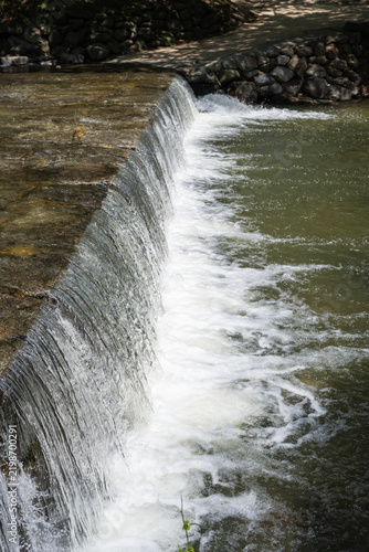 Clear fresh stream water cascading over stone weir creating white foam in peaceful outdoor nature landscape.