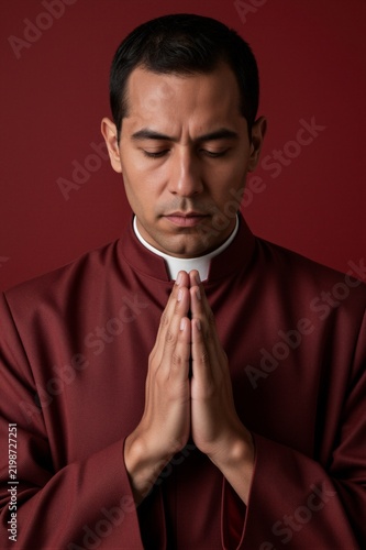 A Catholic priest in a maroon cassock and white clerical collar is posed in prayerful repose against a deep burgundy backdrop.