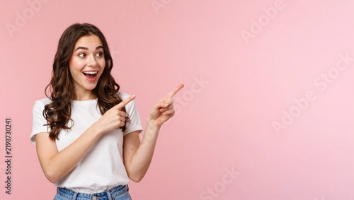 Excited young woman pointing at copy space, surprised female model showing product advertisement, happy girl gesturing towards pink background