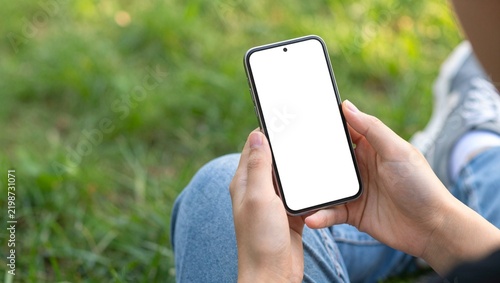 Smartphone mockup with blank white screen, person holding mobile phone in hands outdoors, digital device template for app design on green grass background