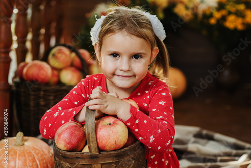 A cute Caucasian five-year-old girl with a basket of apples sits on the porch of her house decorated with pumpkins.