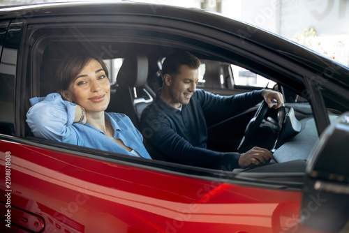 Young couple looking for a new car at a dealership, a woman smiling at the camera sitting inside a modern red vehicle