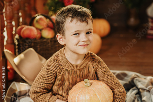 Portrait of a Caucasian boy in a sweater holding a pumpkin on the porch of his house in the autumn season.
