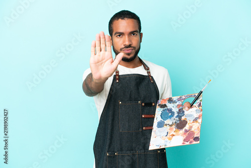 Cuadro en lienzo Young artist latin man holding a palette isolated on blue background making stop