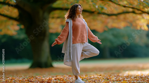 Woman practicing balance beneath a large tree in autumn park, wearing a rust sweater and light trousers among fallen leaves, outdoor wellness portrait scene
