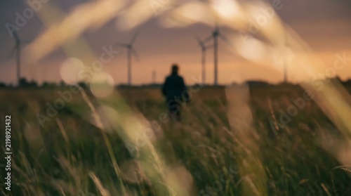 A solitary person walking through tall green grass in a field with wind turbines spinning under sunset sky