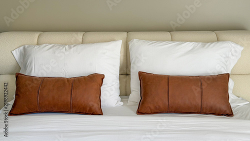 Modern cozy bed with white bedding and brown decorative pillows, neutral bedroom interior with soft upholstered headboard and minimalist hotel style.