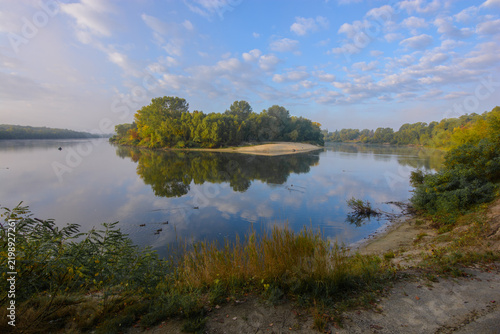 Obraz na plátně A beautiful green island reflects perfectly on the calm, mirror-like surface of a wide river under a bright blue sky