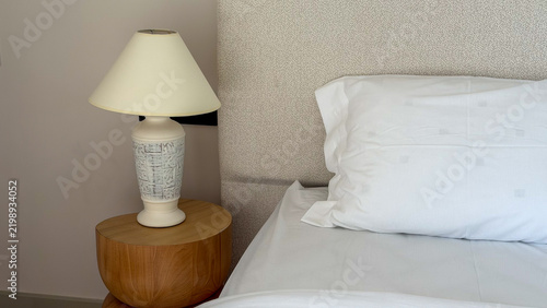 Cozy modern bedroom detail with white bedding, upholstered headboard, wooden bedside table and decorative table lamp in a neutral contemporary interior.