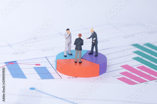Miniature corporate team discussing business statistics on pie chart surrounded by bar charts