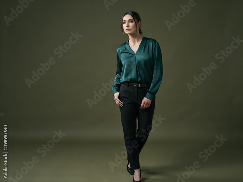 Full body shot of a woman in a dark emerald green satin v-neck blouse and black trousers, walking towards the camera on a grey background